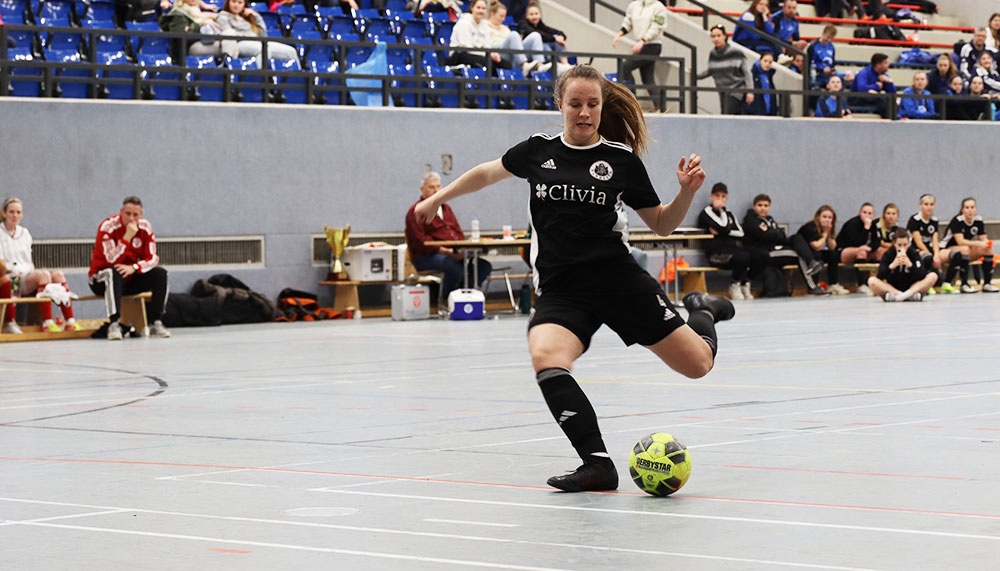 [{"clang_id":1,"value":"Futsalspiel: Spielerin in schwarzem Trikot mit Ball, hochgehobenem Bein. Im Hintergrund Zuschauertribüne und weitere Spieler."}]