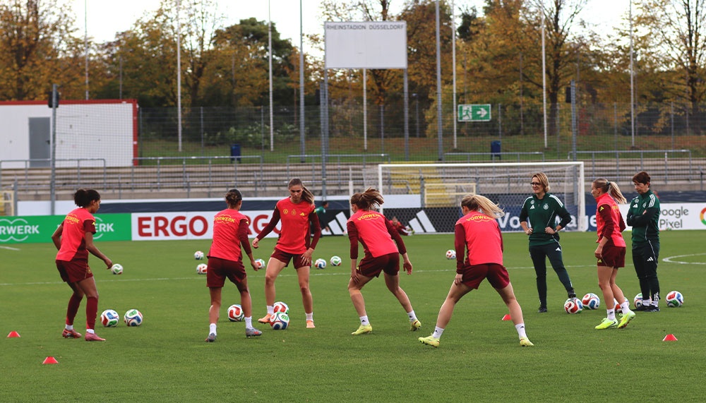 [{"clang_id":1,"value":"Fußballspielerinnen trainieren auf grünem Rasen. Einige dribbeln den Ball, andere stehen bereit. Im Hintergrund Gebäude und Bäume mit Herbstlaub."}]