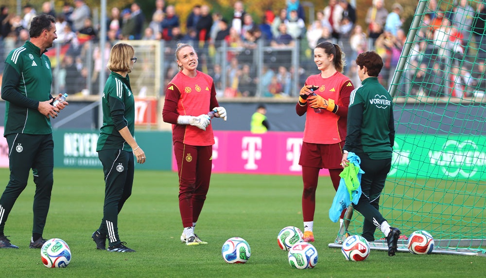 [{"clang_id":1,"value":"Fußballspielerinnen und Trainer stehen auf grünem Rasen vor einem Fußballtor. Einige halten Fußbälle oder Trainingsutensilien. Im Hintergrund Zuschauer."}]