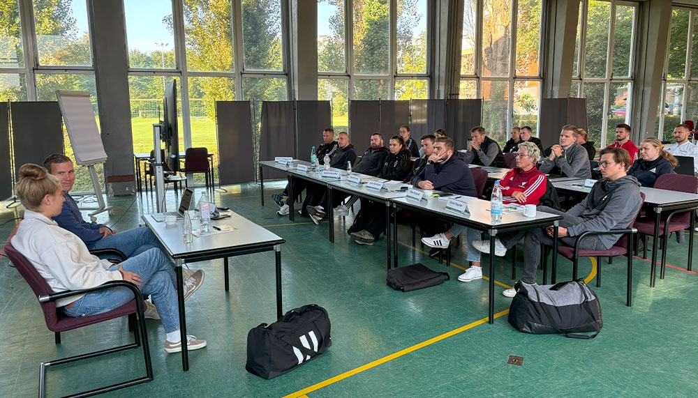 [{"clang_id":1,"value":"Raum mit grossen Fenstern, in dem etwa 20 Personen an einem langen Tisch sitzen. Einige tragen Sportkleidung, vor ihnen stehen Wasserflaschen und Namensschilder. Zwei Personen sitzen seitlich in Stühlen."}]