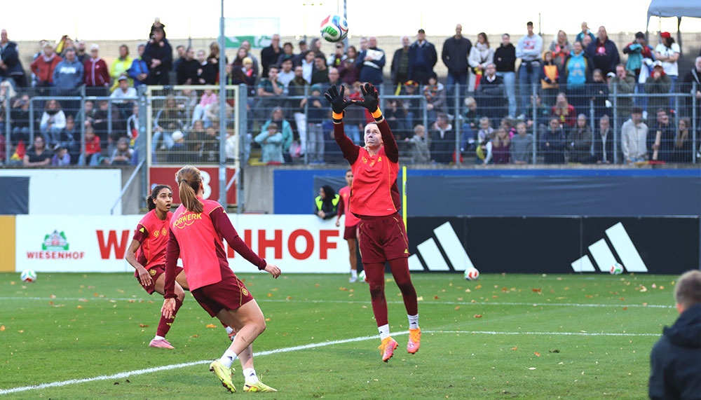 [{"clang_id":1,"value":"Fußballspiel: Torwartin springt hoch, um Ball abzuwehren. Zwei Spielerinnen im Vordergrund, Zuschauerreihen im Hintergrund. Rasenplatz mit Linien."}]