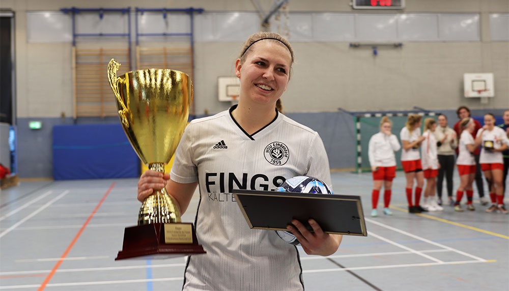 [{"clang_id":1,"value":"Handballspielerin mit zwei Trophäen in einer Sporthalle. Im Hintergrund stehen weitere Spielerinnen."}]