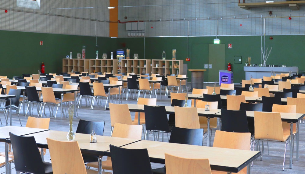 [{"clang_id":1,"value":"Cafeteria mit vielen leeren Tischen und Stühlen. Im Hintergrund eine Theke mit Getränkespendern und Notausgängen an den Wänden."}]