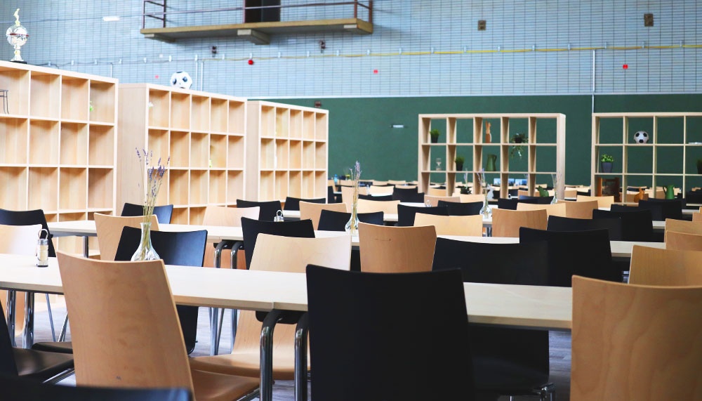 [{"clang_id":1,"value":"Cafeteria oder Mensa mit langen Tischen und Stühlen. Im Hintergrund hohe Regale und eine grüne Wand. Sporthauser Atmosphäre."}]