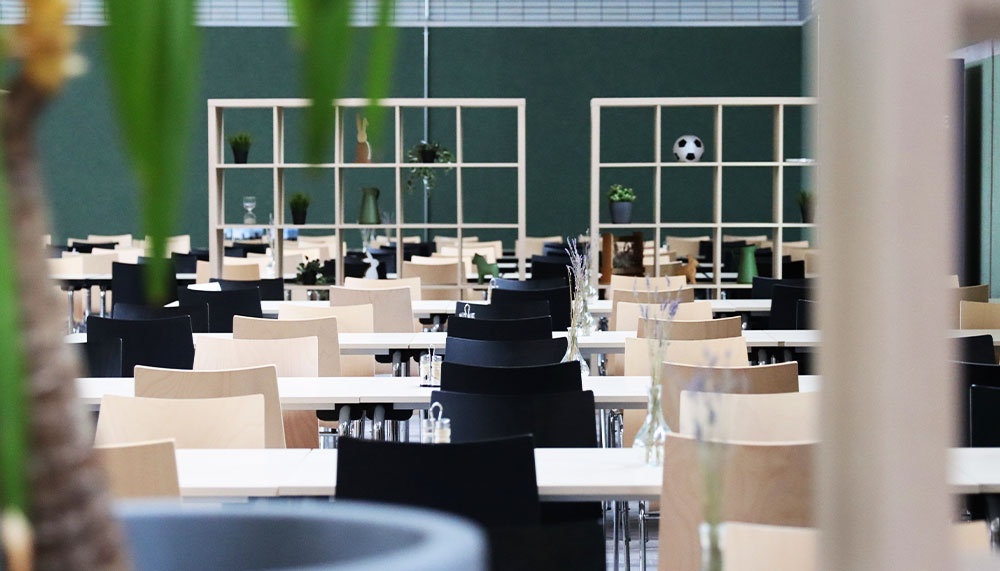 [{"clang_id":1,"value":"Cafeteria oder Mensa mit vielen gedeckten Tischen und Stühlen. Im Hintergrund eine grüne Wand mit weißen Regalen und einem Fußball."}]