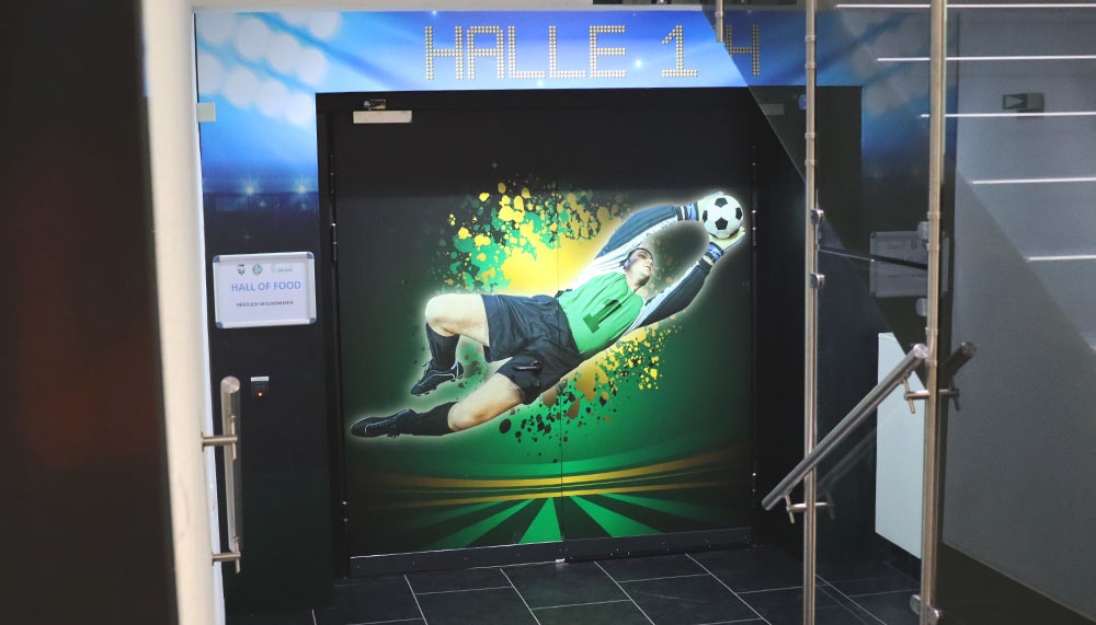 [{"clang_id":1,"value":"Tür mit Fußball-Motiv. Darauf ein Spieler beim Schießen, grüner Hintergrund. Oberhalb der Tür \"HALLE 14\" in blauen Leuchtbuchstaben."}]