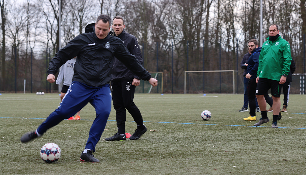 [{"clang_id":1,"value":"Fußballtrainer schießt auf einem Kunstrasenplatz einen Ball. Im Hintergrund weitere Personen und ein Fußballfeld."}]