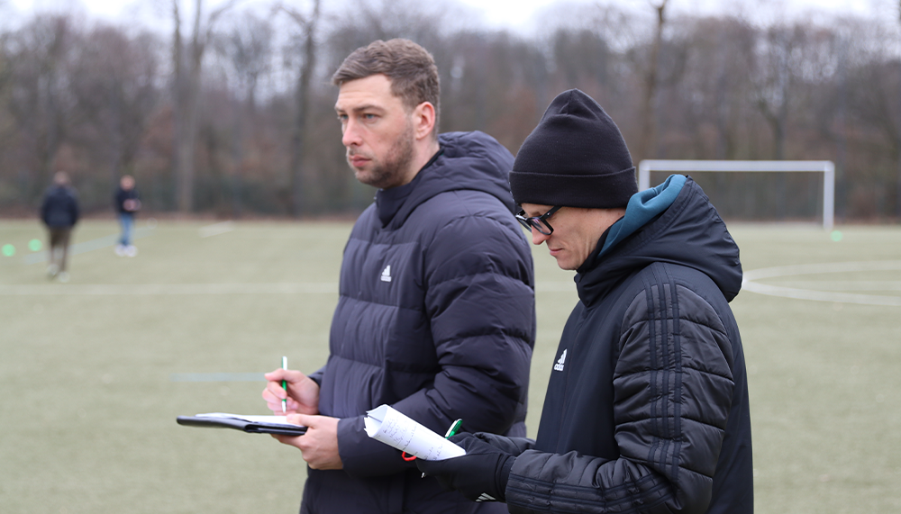 [{"clang_id":1,"value":"Zwei Männer mit Winterkleidung stehen auf einem Fußballfeld. Einer hält ein Clipboard und einen Stift, der andere trägt eine Brille und eine Mütze. Im Hintergrund sind weitere Personen und ein Fußballtor erkennbar."}]