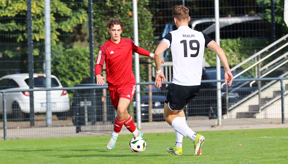 [{"clang_id":1,"value":"Zwei Fußballspieler auf grünem Rasen: Links in rot, dribbelt den Ball; rechts in schwarz-weiß, verfolgt ihn. Im Hintergrund ein Zaun und Fahrzeuge."}]