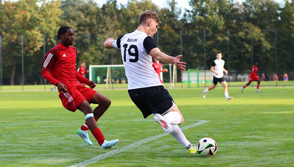 [{"clang_id":1,"value":"Fußballspiel: Spieler mit Nr. 19 schießt Ball, Gegenspieler in Rot versucht Blocken. Grünfläche, Bäume im Hintergrund."}]