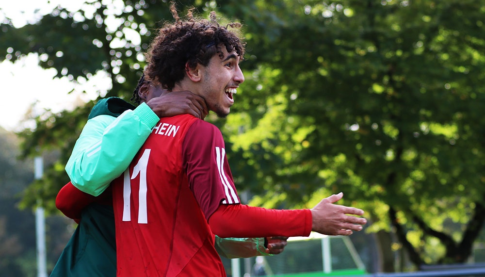 [{"clang_id":1,"value":"Zwei Fußballspieler jubeln eng umschlungen. Der vordere Spieler trägt ein rotes Trikot mit der Nummer 11, der andere ein grünes Trikot mit gelben Ärmeln. Im Hintergrund Bäume."}]