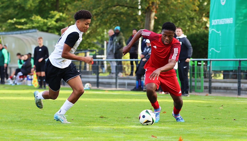 [{"clang_id":1,"value":"Zwei junge Fußballspieler auf grünem Rasen. Links trägt ein Spieler ein weiß-schwarzes Trikot, rechts ein rotes. Beide dribbeln den Ball. Im Hintergrund Zuschauer und Bäume."}]