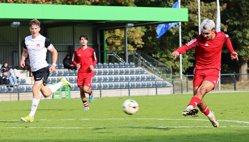 [{"clang_id":1,"value":"Fußballspiel: Ein Spieler in Rot schießt auf das Tor, während zwei weitere Spieler in Weiß und Rot ihm entgegenlaufen. Hintergrund: Stadiontribüne und Bäume."}]