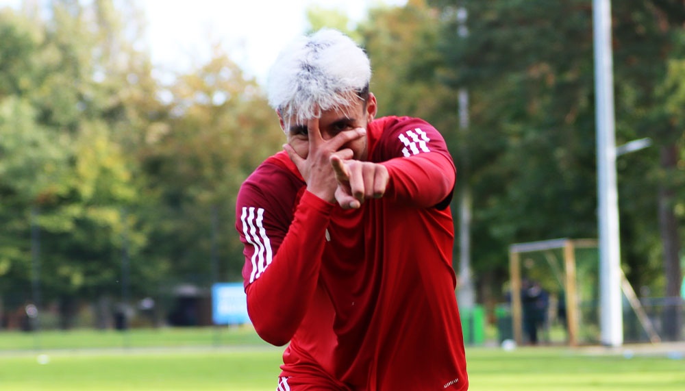 [{"clang_id":1,"value":"Fußballspieler mit weiß gefärbten Haaren in rotem Trainingsanzug, Arme angewinkelt, blickt entschlossen in die Kamera. Hintergrund: Fußballfeld mit Bäumen."}]