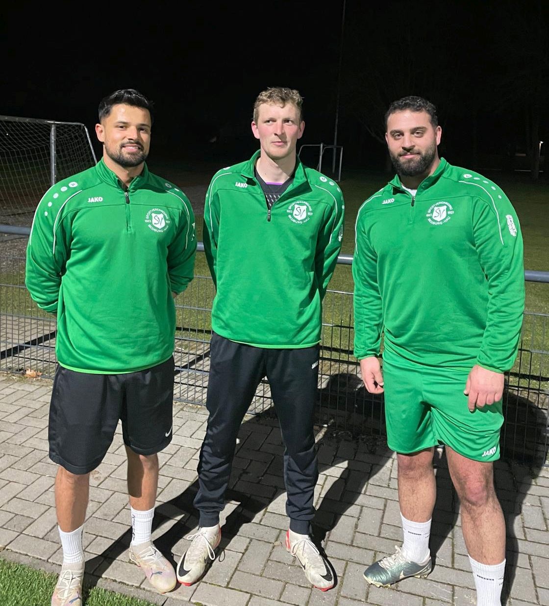 [{"clang_id":1,"value":"Drei Männer in grünen Trainingsanzügen und Shorts stehen auf einem Fußballplatz vor einem dunklen Hintergrund."}]
