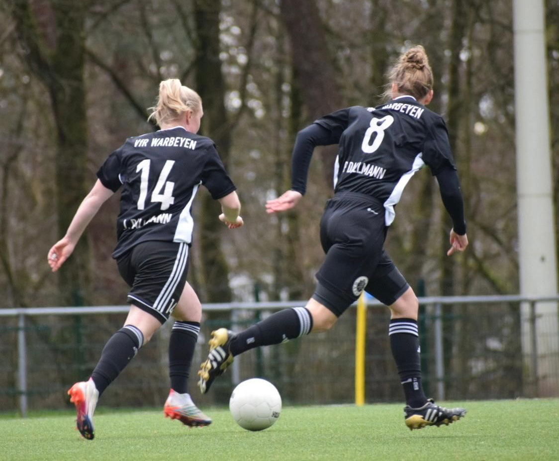 [{"clang_id":1,"value":"Zwei Fußballspielerinnen mit blonden Zöpfen in schwarzen Trikots mit den Nummern 14 und 8 spielen auf einem grünen Rasenplatz. Eine Spielerin schießt auf den Ball."}]