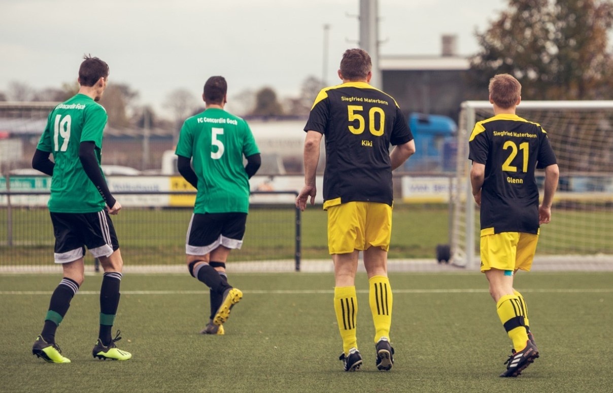 [{"clang_id":1,"value":"Vier Fußballspieler mit Rückenansicht auf einem grünen Rasenplatz. Spieler tragen Trikots mit den Nummern 19, 5, 50 und 21. Im Hintergrund ein Fußballtor und Gebäude."}]