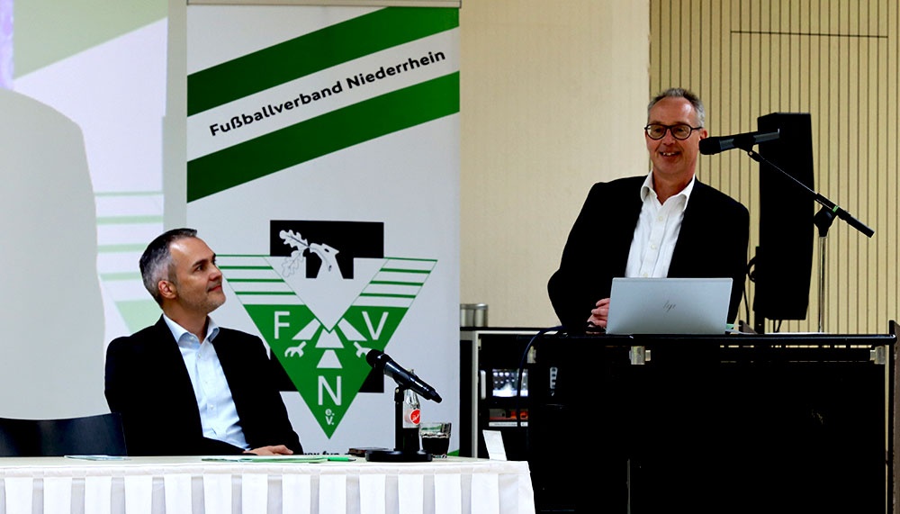 [{"clang_id":1,"value":"Zwei Männer an einem Tisch vor einem Banner mit Fußballverein-Logo \"Fußballverband Niederrhein\". Der Mann rechts spricht in ein Mikrofon."}]