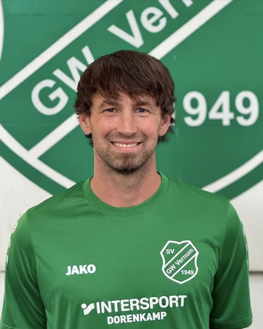 [{"clang_id":1,"value":"Mann mit kurzbraunen Haaren lächelt in die Kamera. Er trägt ein grünes Sporttrikot mit dem Logo \"SV GW Vernum 1949\" und Werbeaufdrucken. Im Hintergrund eine grüne Fläche mit Aufschrift."}]