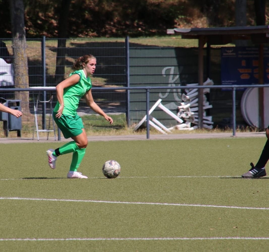 [{"clang_id":1,"value":"Fußballspiel: Junge Spielerin in grünem Trikot dribbelt den Ball auf einem grünen Kunstrasenplatz. Im Hintergrund eine Zaun- und Gebäudekulisse."}]