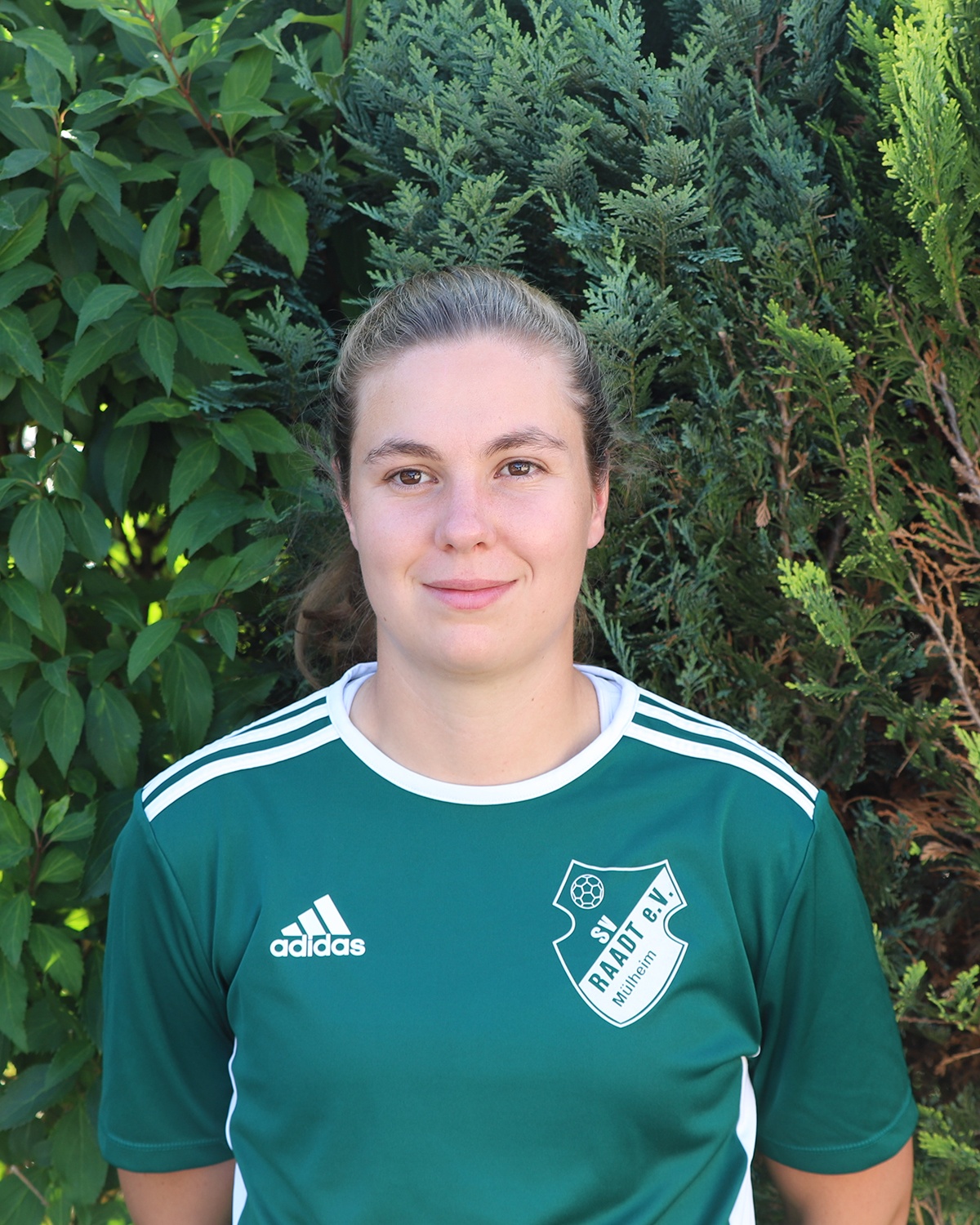 [{"clang_id":1,"value":"Junge Frau mit braunen Haaren trägt grünes Fußballtrikot mit weißem Logo \"ST Raadt e.V. Mülheim\" und Adidas-Logo. Im Hintergrund grüner Strauch."}]