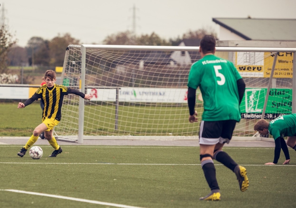 [{"clang_id":1,"value":"Jugendfußballspiel: Spieler in Gelb schießt aufs Tor, verteidigt von grün gekleideten Spielern auf grünem Rasenfeld. Im Hintergrund Gebäude und Werbebanner."}]