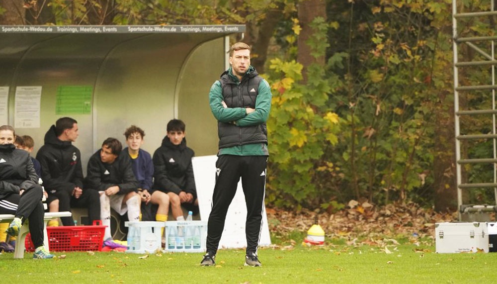 [{"clang_id":1,"value":"Fußballtrainer mit verschränkten Armen vor einer Bank, auf der Fußballspieler sitzen. Im Hintergrund ein Wald."}]