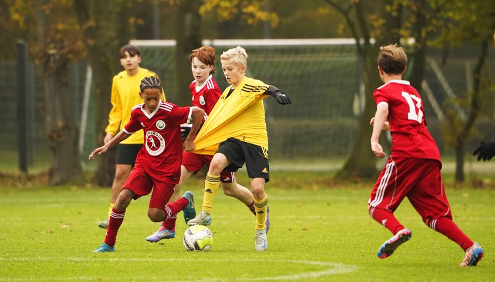 [{"clang_id":1,"value":"Jugendfußballspiel: Spieler in roten und gelben Trikots kämpfen um den Ball auf grünem Rasen. Im Hintergrund weitere Spieler und Bäume."}]