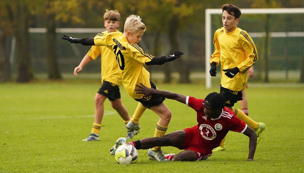 [{"clang_id":1,"value":"Fußballspiel: Zwei jugendliche Spieler kämpfen um den Ball. Ein Spieler in Rot rutscht, während ein Spieler in Gelb versucht, den Ball zu erobern. Im Hintergrund weitere Spieler und ein Fußballtor."}]