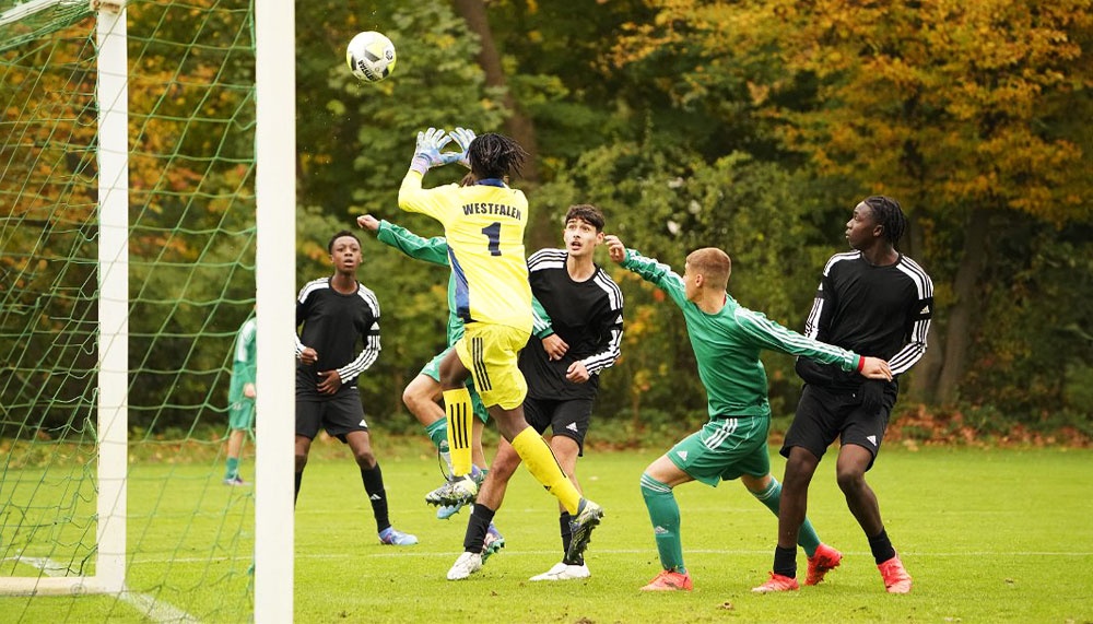 [{"clang_id":1,"value":"Fußballspiel: Torwart springt hoch, um Ball abzuwehren. Mehrere Spieler um ihn herum, grünes Spielfeld, Tornetz im Hintergrund."}]