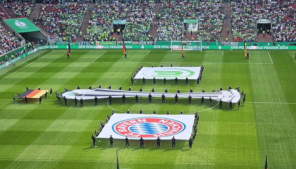 [{"clang_id":1,"value":"Fußballstadion, gefüllt mit Zuschauern. Auf dem Rasen eine große Bayern München Vereinsflagge und Personen mit Flaggen, vermutlich vor einem Spiel."}]