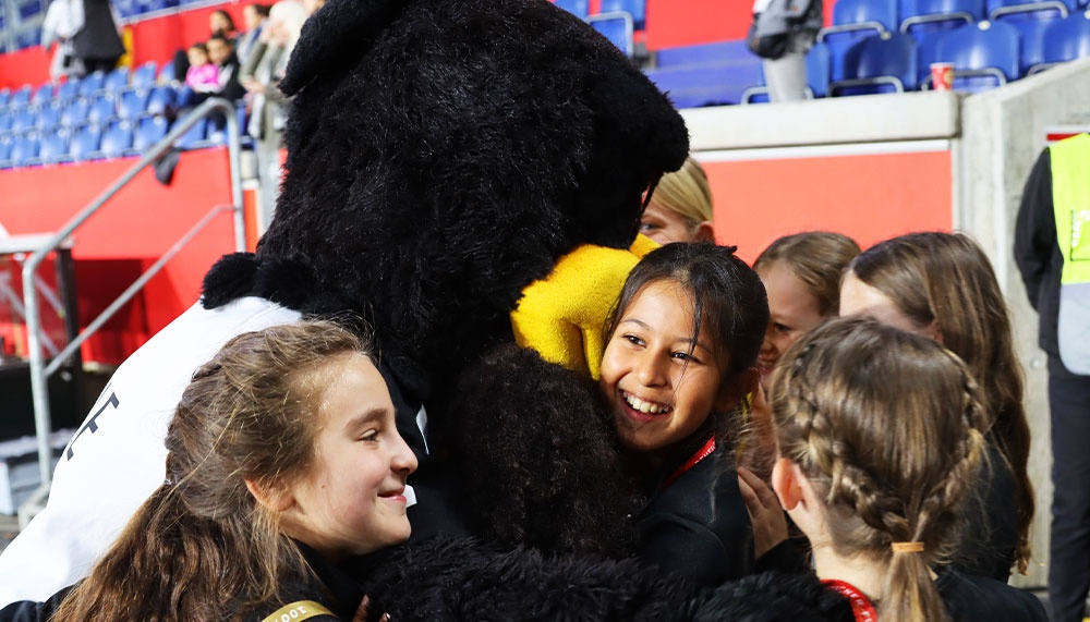 [{"clang_id":1,"value":"Mascot im Tierkostüm umarmt lächelnde Kinder in einem Stadion. Im Hintergrund sind leere Sitze und Zuschauerreihen erkennbar."}]