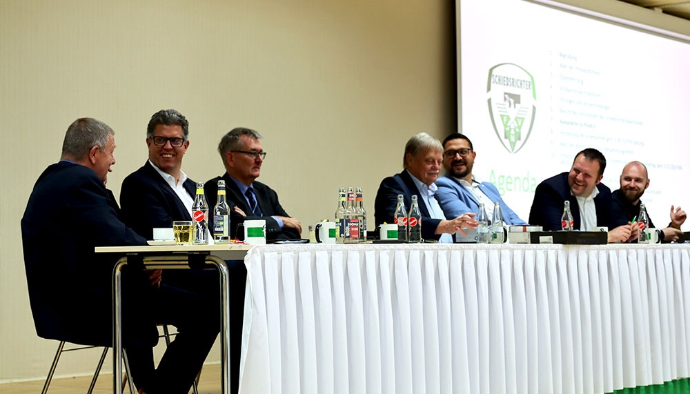[{"clang_id":1,"value":"Konferenzteilnehmer sitzen an einem Tisch vor einer Leinwand mit Agenda. Sie tragen Anzüge und Hemden. Auf dem Tisch stehen Wassergläser und Notizblöcke."}]