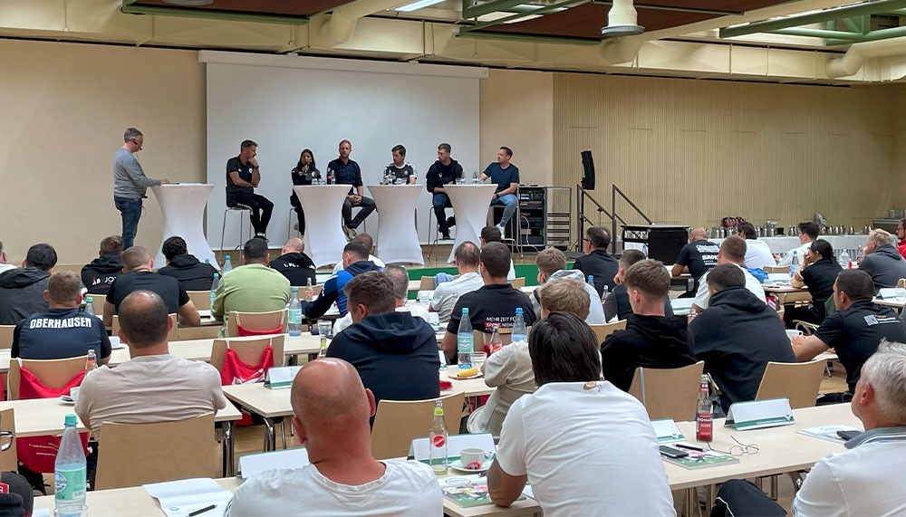 [{"clang_id":1,"value":"Großer Konferenzraum mit Publikum auf Stühlen vor einer Bühne. Auf der Bühne sitzen Personen an einem Tisch, vor dem ein Redner steht."}]