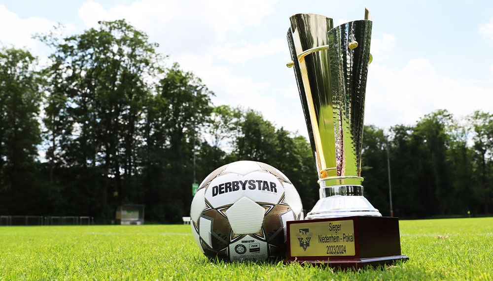 [{"clang_id":1,"value":"Fußballpokal \"Niederhein-Pokal 2023/2024\" und Derby Star Fußball auf grüner Wiese vor Baumkulisse und bewölktem Himmel."}]