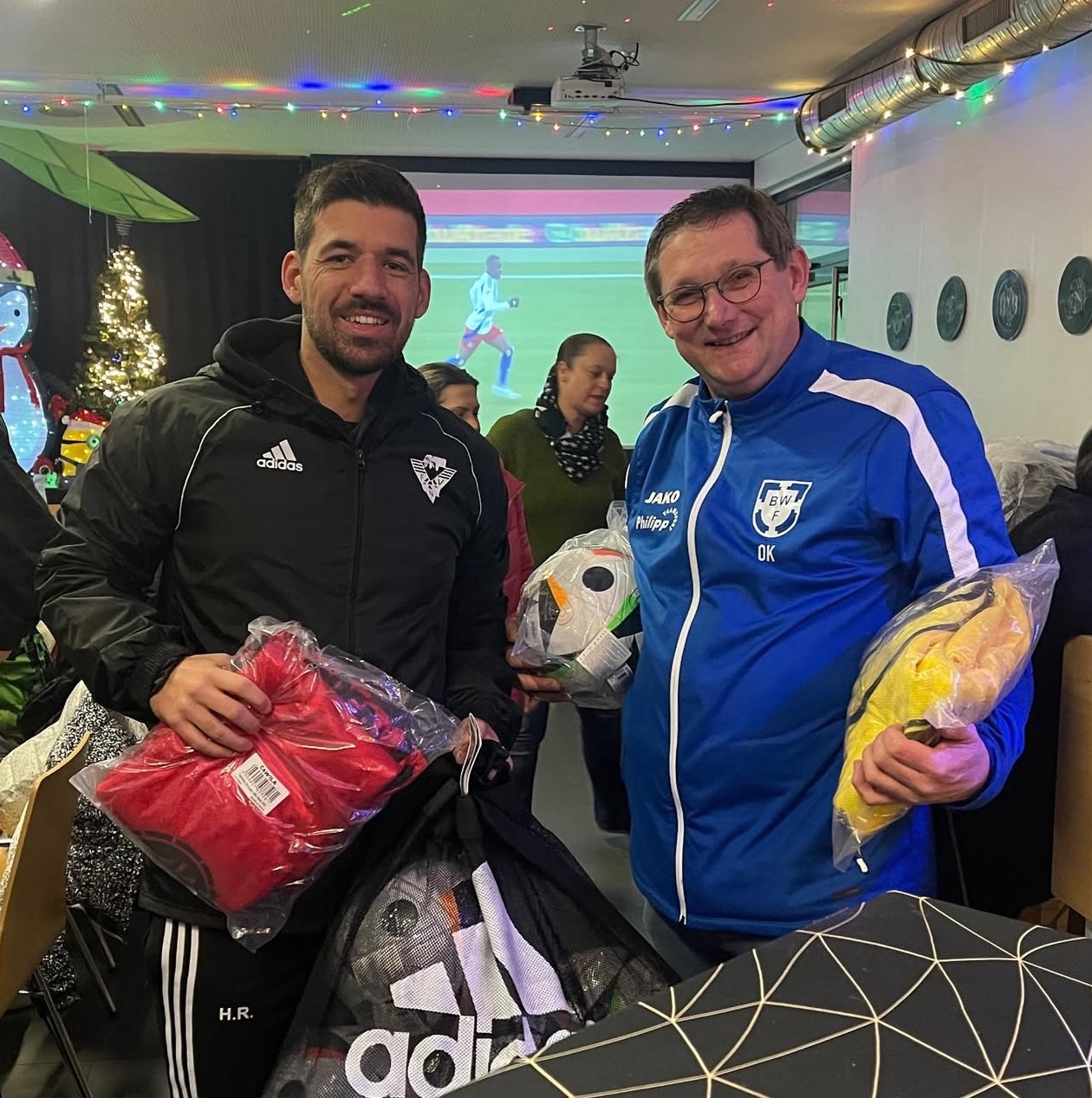 [{"clang_id":1,"value":"Zwei Männer lächeln in die Kamera. Links trägt ein Mann dunkle Sportkleidung und hält rote Geschenke, rechts ein Mann blaue Sportkleidung mit gelben Geschenken. Im Hintergrund sind weitere Personen und Weihnachtsdekoration zu sehen."}]