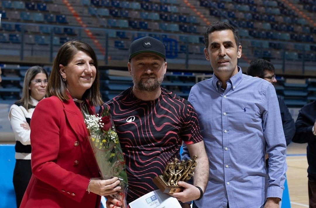 [{"clang_id":1,"value":"Drei Personen stehen in einer Sporthalle: eine Frau links mit Blumen, in der Mitte ein Mann mit Kappe und Preis, rechts ein Mann im Hemd. Im Hintergrund Zuschauerreihen."}]
