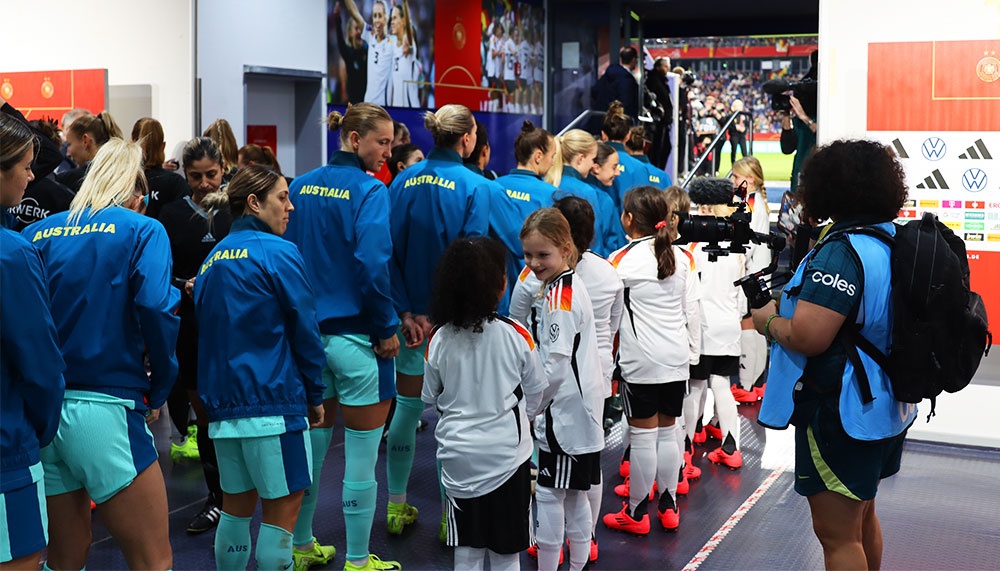 [{"clang_id":1,"value":"Fußballspielerinnen der Teams Australien (blau) und Spanien (weiß) stehen im Tunnel eines Stadions. Im Hintergrund Zuschauer und Kameras."}]