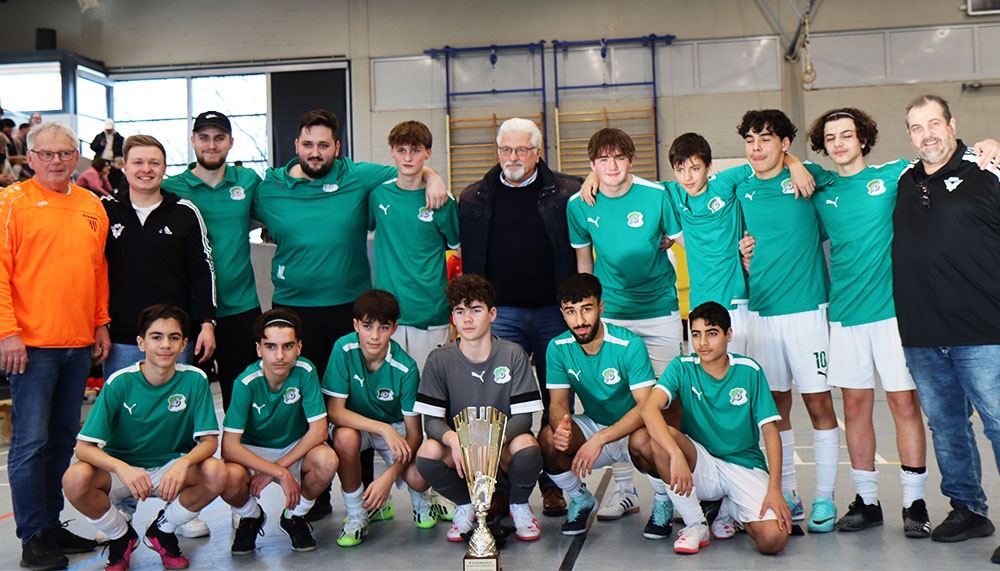 [{"clang_id":1,"value":"Fußballmannschaft in grünen Trikots und weißen Shorts posiert mit goldenem Pokal in einer Sporthalle. Einige Betreuer stehen daneben."}]