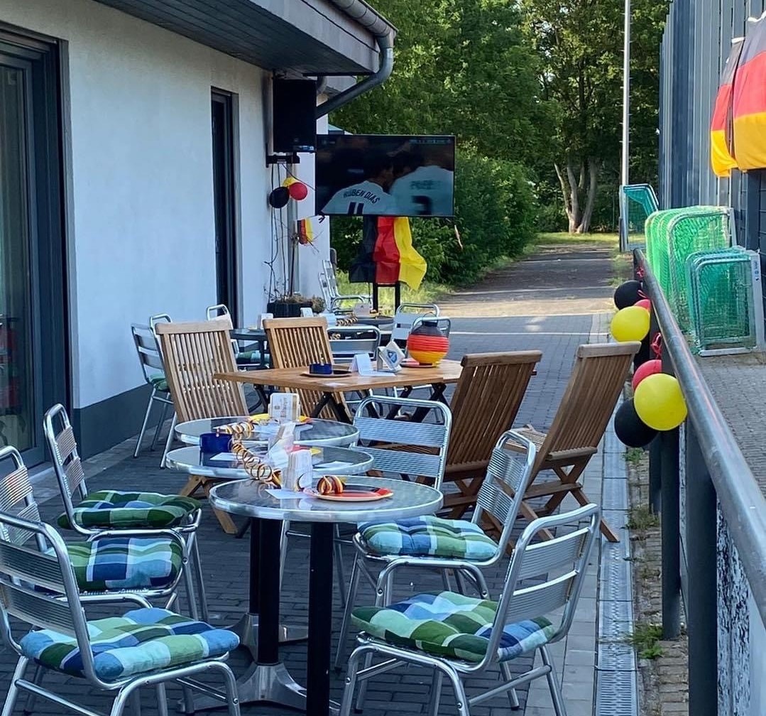 [{"clang_id":1,"value":"Terrasse eines Cafés oder Restaurants. Tische und Stühle mit blau-grünen Kissen stehen auf gepflastertem Untergrund. Im Hintergrund sind deutsche Flaggen und Dekorationen zu sehen."}]