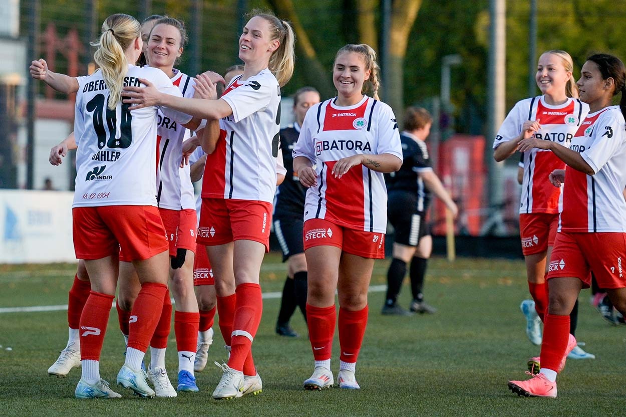 [{"clang_id":1,"value":"Frauenfußballteam jubelt auf dem Spielfeld. Spielerinnen in roten und weißen Trikots umarmen sich. Hintergrund: Zuschauer und Bäume."}]