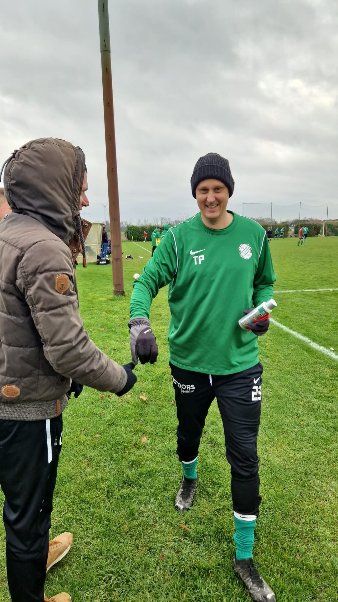 [{"clang_id":1,"value":"Zwei Männer auf Grünfläche, einer mit Hoody und brauner Jacke, der andere in grünem Trainingsanzug und schwarzer Mütze. Im Hintergrund weitere Personen und ein Mast."}]