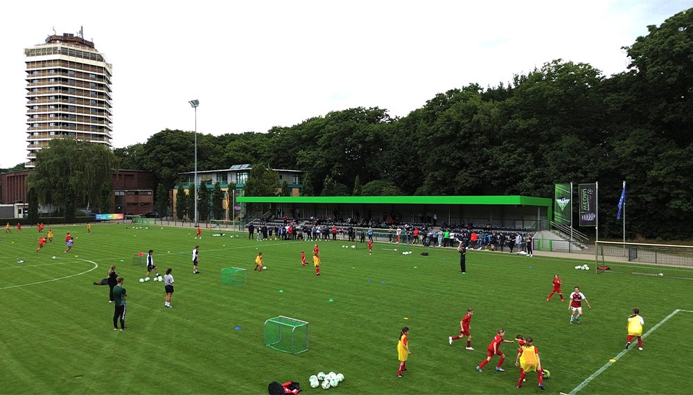 [{"clang_id":1,"value":"Fußballfeld mit trainierenden Kindern und Jugendlichen. Im Hintergrund ein Hochhaus und Bäume unter grauem Himmel."}]