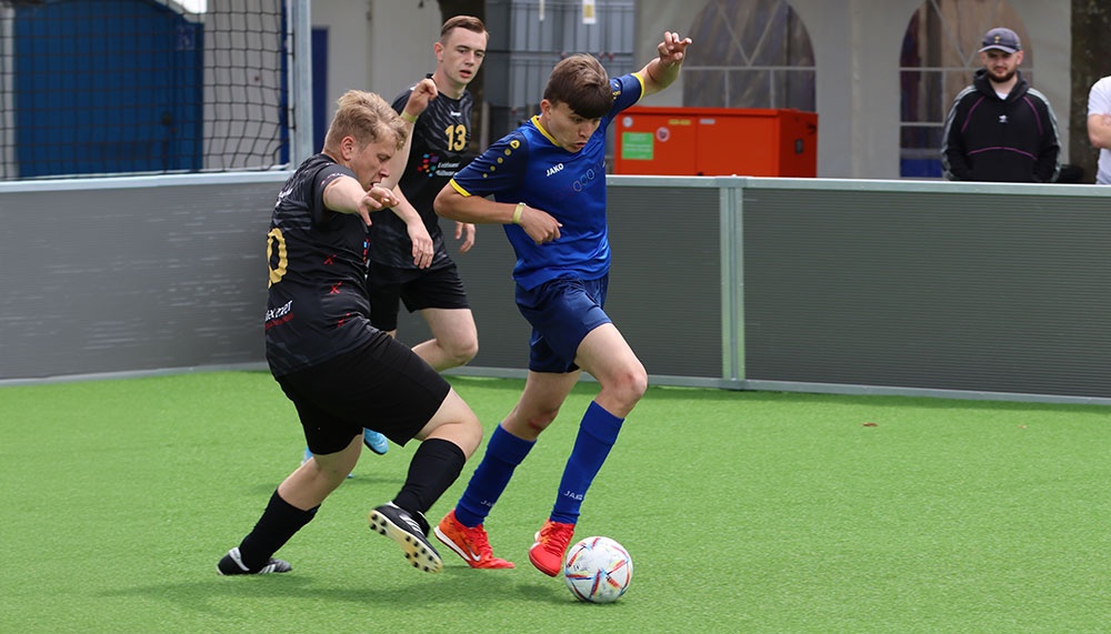 [{"clang_id":1,"value":"Drei Fussballspieler auf grünem Kunstrasen. Ein Spieler in Blau mit rotem Schuhwerk dribbelt den Ball, während zwei Gegner ihn verfolgen. Im Hintergrund Zuschauer und eine Metallbarriere."}]