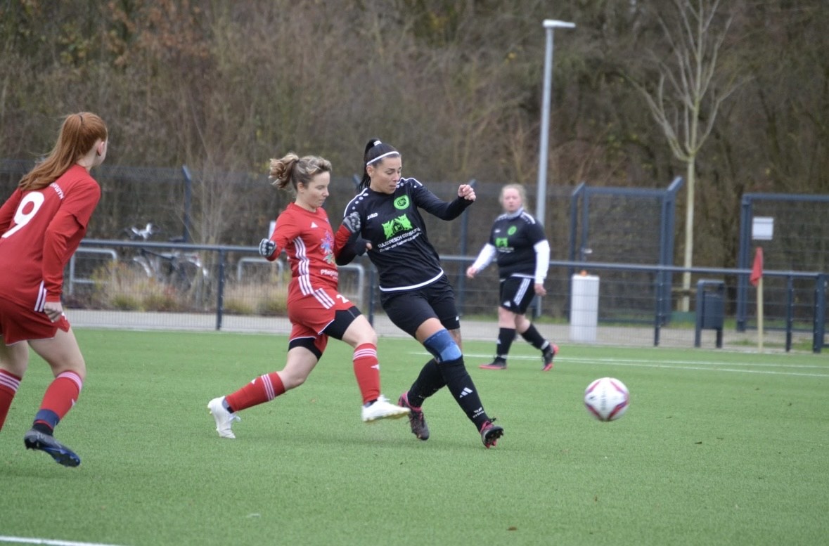 [{"clang_id":1,"value":"Fußballspiel. Drei Spielerinnen in Aktion auf grünem Rasen. Eine Spielerin in Schwarz schießt den Ball, zwei Gegenspielerinnen in Rot stehen in der Nähe. Im Hintergrund Bäume und eine Zuschauertribüne."}]