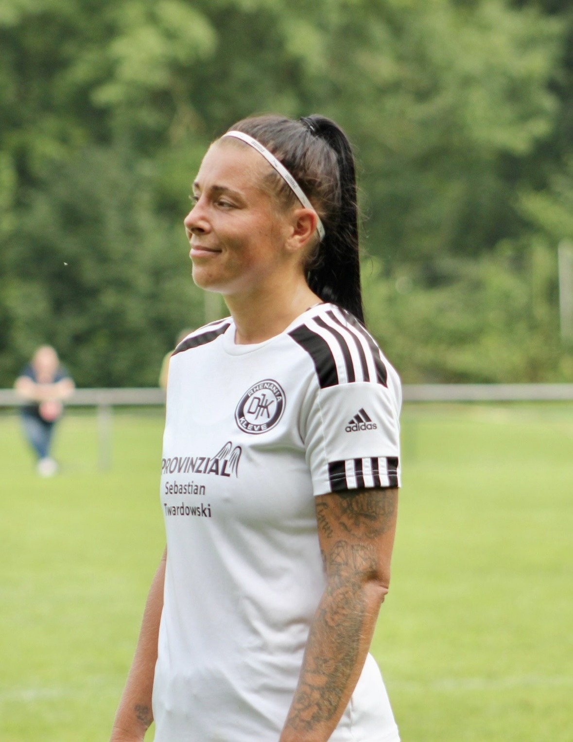 [{"clang_id":1,"value":"Fußballspielerin mit Stirnband und Trikot des Vereins „DJK Schwarz-Weiß“ steht auf einem grünen Rasenfeld. Sichtbare Tattoos am Arm."}]