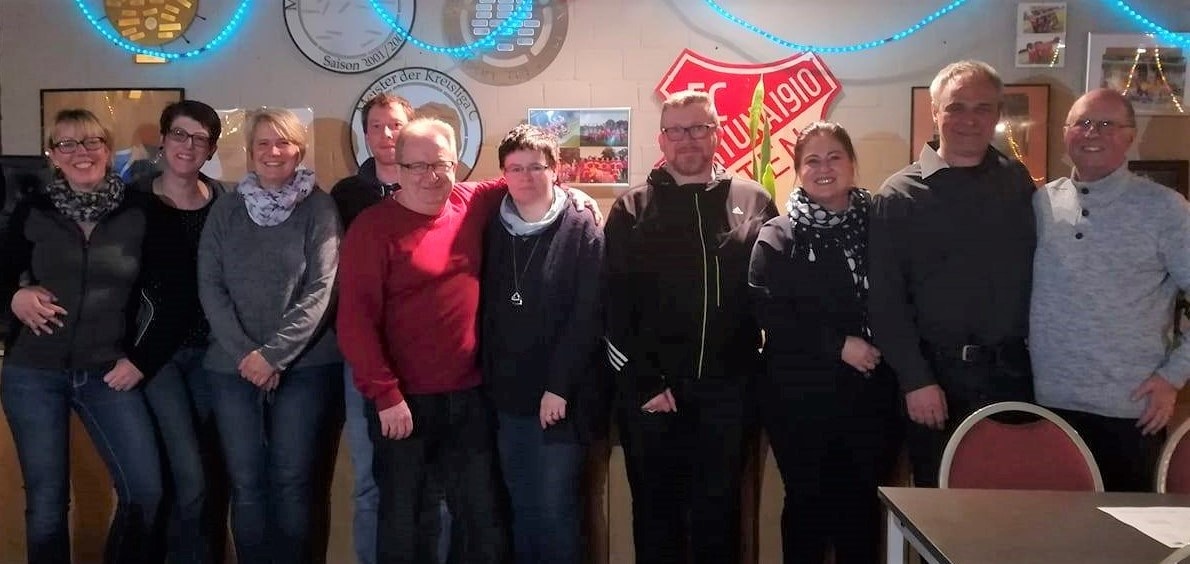 [{"clang_id":1,"value":"Gruppe von neun Personen steht lächelnd vor einer Wand mit Fußball-Emblemen und Dekoration. Unterschiedliche Kleidung, überwiegend Freizeitkleidung."}]