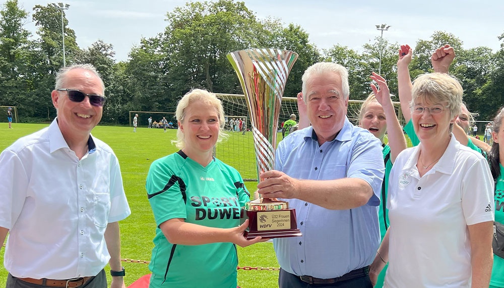 [{"clang_id":1,"value":"Vier Personen posieren mit einem silbernen Pokal auf einem Fußballfeld. Links und rechts weiße Hemden, in der Mitte ein grünes Fußballtrikot. Im Hintergrund Spielende und Zuschauer."}]