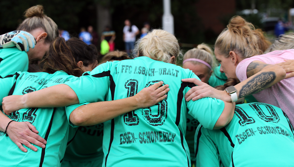 [{"clang_id":1,"value":"Frauenfußballteam im Kreis, Arme um Schultern. Alle tragen türkisfarbene Trikots mit dem Schriftzug \"1. FFC Asbach-Uralt Essen-Schonnebeck\" und Nummern."}]