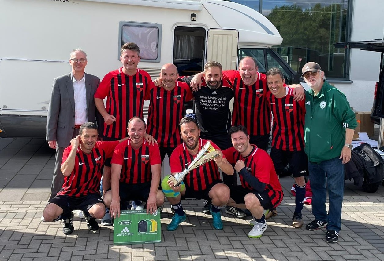 [{"clang_id":1,"value":"Fußballmannschaft in rot-schwarzen Trikots posiert mit Pokal vor einem weißen Wohnmobil. Mehrere Männer stehen oder knien davor."}]